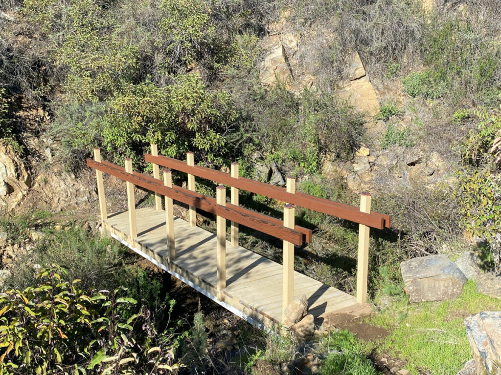small wooden footbridge with railings, leading across a stream bed and surrounded by rocks and shrubs.