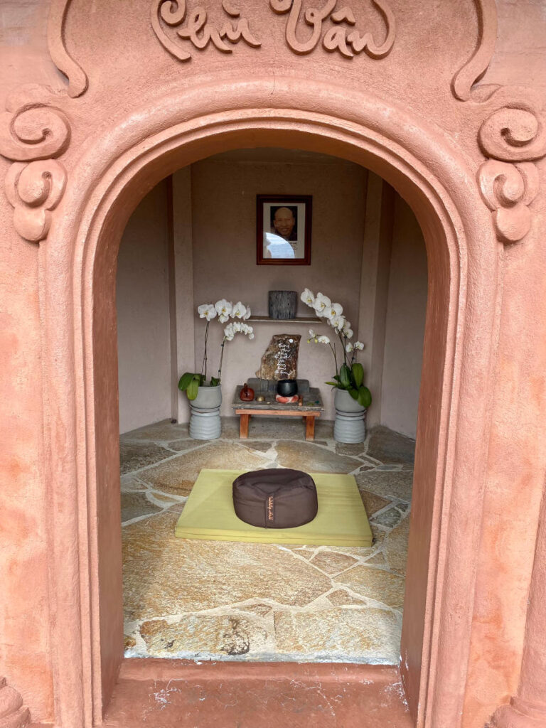 the clay-colored, rounded doorway of the stupa, leading into a small room with a mat and cushion in front of a small altar with flowers, bells, and a framed photo of a monk on the wall.